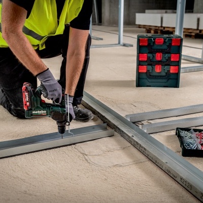 Homem a usar ferramenta elétrica para montar estrutura metálica em chão de cimento com caixas de ferramentas próximas.