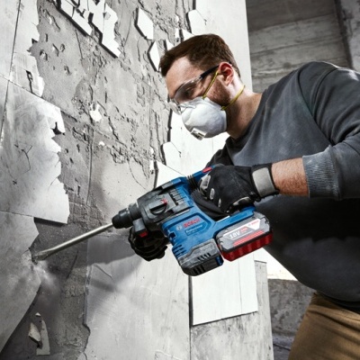 Homem com máscara e óculos de segurança perfura parede cinza com furadeira Bosch azul e vermelha