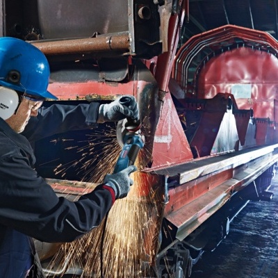 Operário industrial com capacete azul e protetores auriculares a usar esmerilhadora elétrica em equipamento metálico vermelho