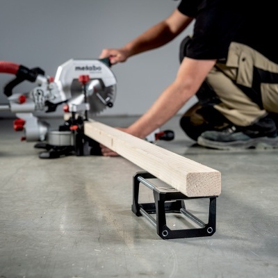 Serra de cortar madeira Metabo com cabo vermelho e punho branco, tábua de madeira clara em suporte preto sobre chão cinza.