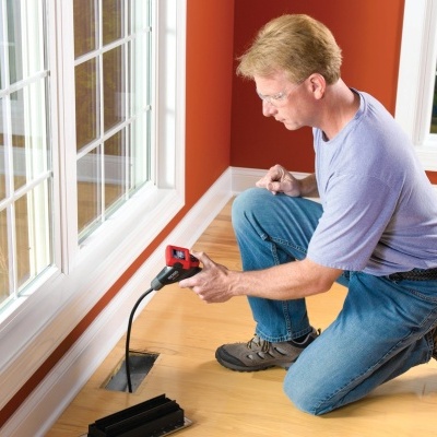 Homem a usar aparelho de inspeção preto e vermelho junto a parede branca e chão de madeira
