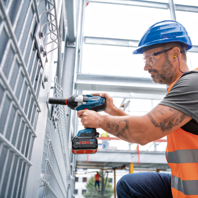 Trabalhador com colete e capacete a usar furadeira numa estrutura metálica num ambiente industrial