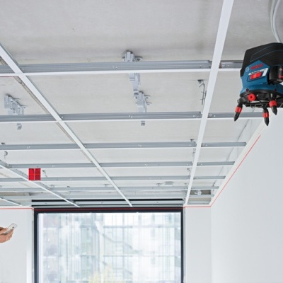 Nível a laser BOSCH azul e preto fixado em parede branca com linhas vermelhas, homem ao lado segurando controle