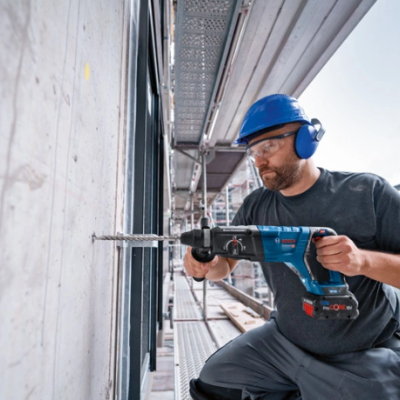 Homem com capacete e proteção a usar furadeira elétrica numa parede clara em obra.