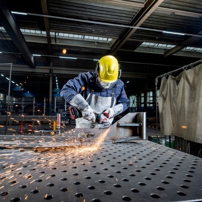 Trabalhador com capacete amarelo e viseira a lixar chapa metálica em oficina