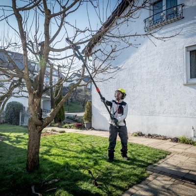 Homem a podar árvore com ferramenta elétrica em jardim junto a casa branca