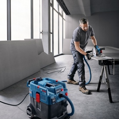 Homem usando ferramenta de corte elétrica ligada a aspirador industrial Bosch azul e preto num espaço industrial