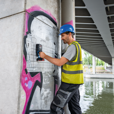 Homem com capacete azul e colete amarelo usando ferramenta Bosch numa coluna de betão com graffiti e grelha de 3D Imaging.