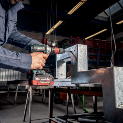 Parafusadeira Metabo a bateria em uso numa oficina industrial com estrutura metálica prateada.