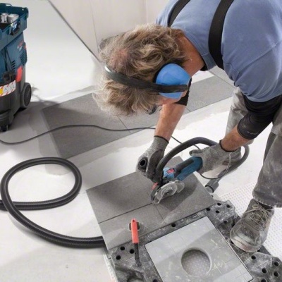 Homem a cortar painel cinzento com ferramenta elétrica azul, máquina azul ao lado, superfície clara.