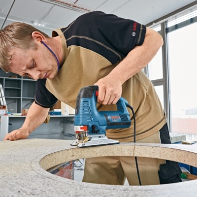 Homem a cortar placa de pedra com serra tico-tico azul numa bancada de trabalho