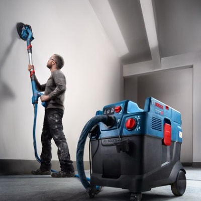 Homem a usar lixadora de parede elétrica azul ligada a máquina de aspiração Bosch
