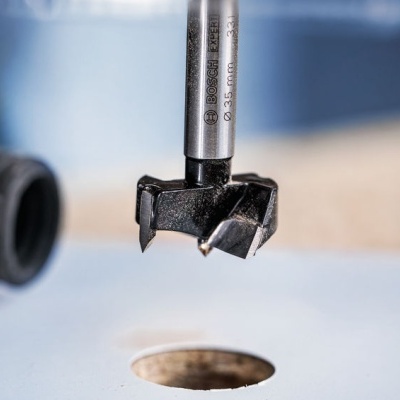 Broca Forstner metálica com parte cortante preta sobre superfície clara com furo