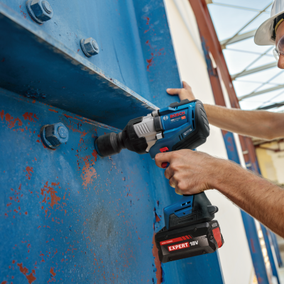 Máquina de impacto BOSCH PRO a bateria com bateria EXPERT 18V em uso numa estrutura metálica azul enferrujada num ambiente industrial.
