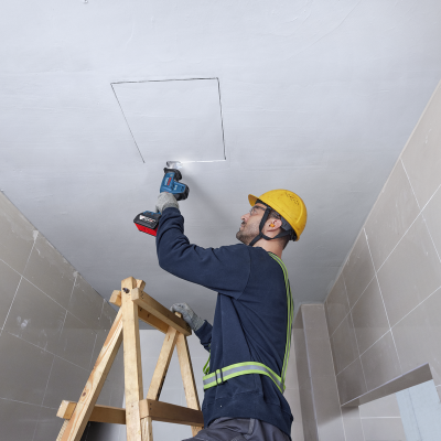 Homem com capacete amarelo em escada usando ferramenta elétrica para trabalhar no teto branco