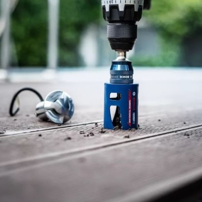 Furadeira Bosch a perfurar madeira com broca azul e texto visível, luva prateada ao lado, ambiente exterior