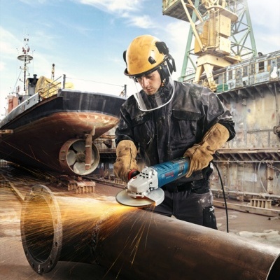 Trabalhador com rebarbadora cortando tubo metálico num estaleiro naval