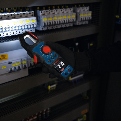 Multímetro digital BOSCH a ser segurado junto a painel elétrico com disjuntores brancos e etiquetas amarelas