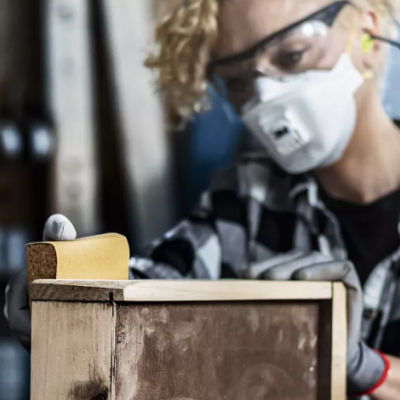 Pessoa a lixar móvel de madeira com máscara e óculos de proteção numa oficina