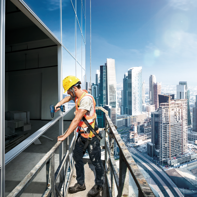 Trabalhador da construção em andaime com furadeira durante construção com arranha-céus ao fundo