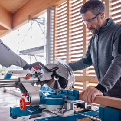 Serra de esquadria Bosch azul e cinza operada por homem com óculos de proteção numa construção de madeira