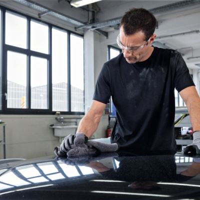 Homem limpa capô de carro preto numa garagem com janelas grandes