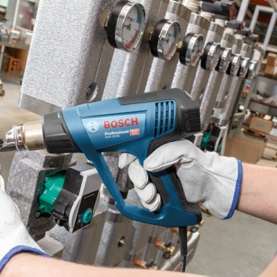 Ferramenta elétrica Bosch Professional GHG 20-63 numa mão com luva branca num ambiente de oficina industrial