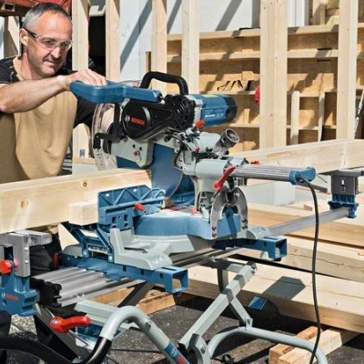 Homem a usar serra de esquadria elétrica azul e preta numa obra com madeira ao fundo