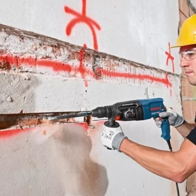 Homem com capacete amarelo a usar ferramenta elétrica Bosch para perfurar parede branca.