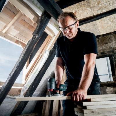 Homem a usar berbequim elétrica numa janela de madeira num sótão com vigas e paredes de pedra