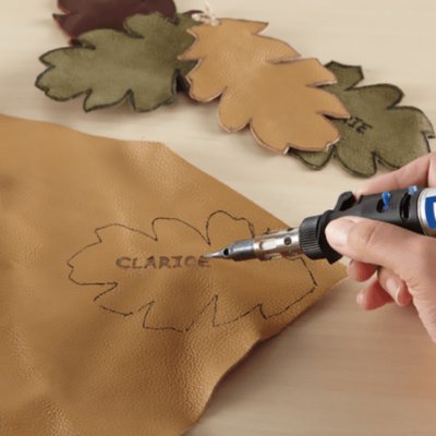 Ferramenta DREMEL gravando pele castanha com molde de folha e texto CLARICE