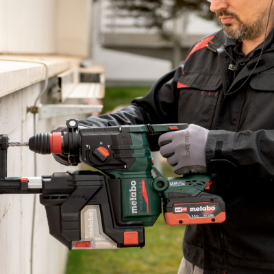 Furadeira sem fio Metabo com coletor de pó e bateria laranja, usada por pessoa com luvas e casaco preto