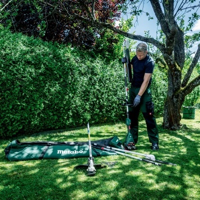Pessoa a usar ferramenta de jardim Metabo em relvado verde com árvores e sebes