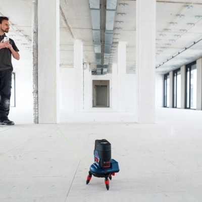 Ferramenta laser preta e azul com tripé vermelho no chão de sala ampla branca, homem observando ao fundo
