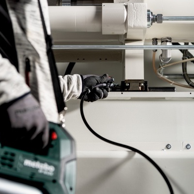 Pessoa a usar ferramenta elétrica num ambiente industrial com tubagens e painéis metálicos.