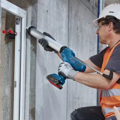 Trabalhador em construção com ferramenta azul e colete laranja a operar na parede de betão