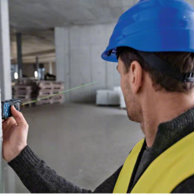 Homem com capacete azul e colete amarelo usa medidor de distância a laser em obra