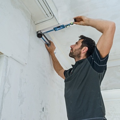 Homem a medir ou ajustar unidade de ar condicionado numa parede branca.