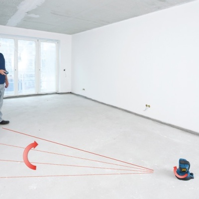 Sala vazia com chão de cimento, paredes brancas, janela e equipamento de medição a laser no chão