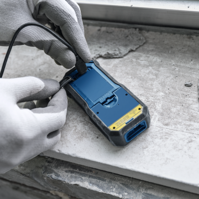 Dispositivo eletrônico azul e preto com etiqueta amarela sendo conectado a um cabo, mãos com luvas cinza, superfície de construção cinzenta com detritos