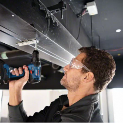 Homem a usar berbequim elétrico azul num teto preto com óculos de proteção.