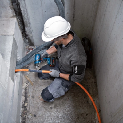 Homem com capacete branco usando furadeira azul em espaço de construção estreito.