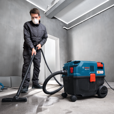Aspirador industrial Bosch PRO azul e preto em uso por trabalhador com máscara, roupa preta e óculos.
