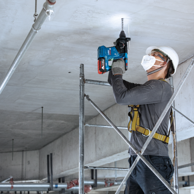 Trabalhador com capacete e máscara usando furadeira Bosch azul para perfurar teto de cimento