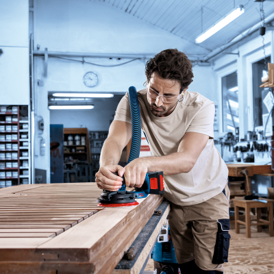 Homem a lixar madeira clara com lixadora elétrica numa oficina de carpintaria