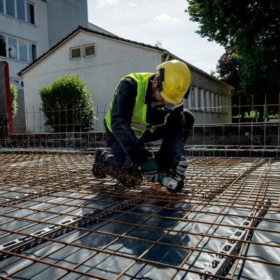 Trabalhador de construção a cortar malha metálica num chão, com capacete amarelo e colete refletor