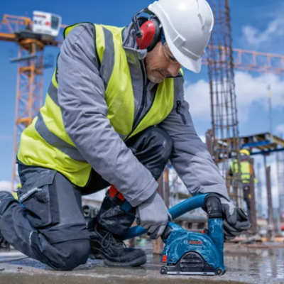 Trabalhador em construção usando ferramenta elétrica azul com capacete branco e colete amarelo