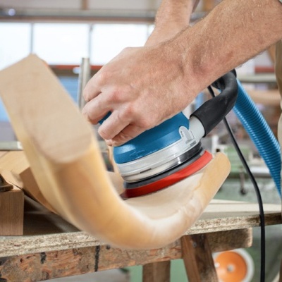 Pessoa polindo pedaço de madeira curvada com ferramenta elétrica numa oficina