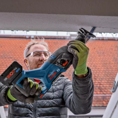 Homem a usar ferramenta eléctrica Bosch azul e preta perto de janela aberta com telhado vermelho ao fundo.