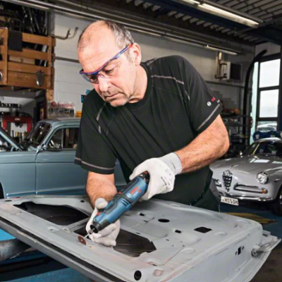 Homem a trabalhar com ferramenta eléctrica em painel metálico num espaço industrial com carros clássicos